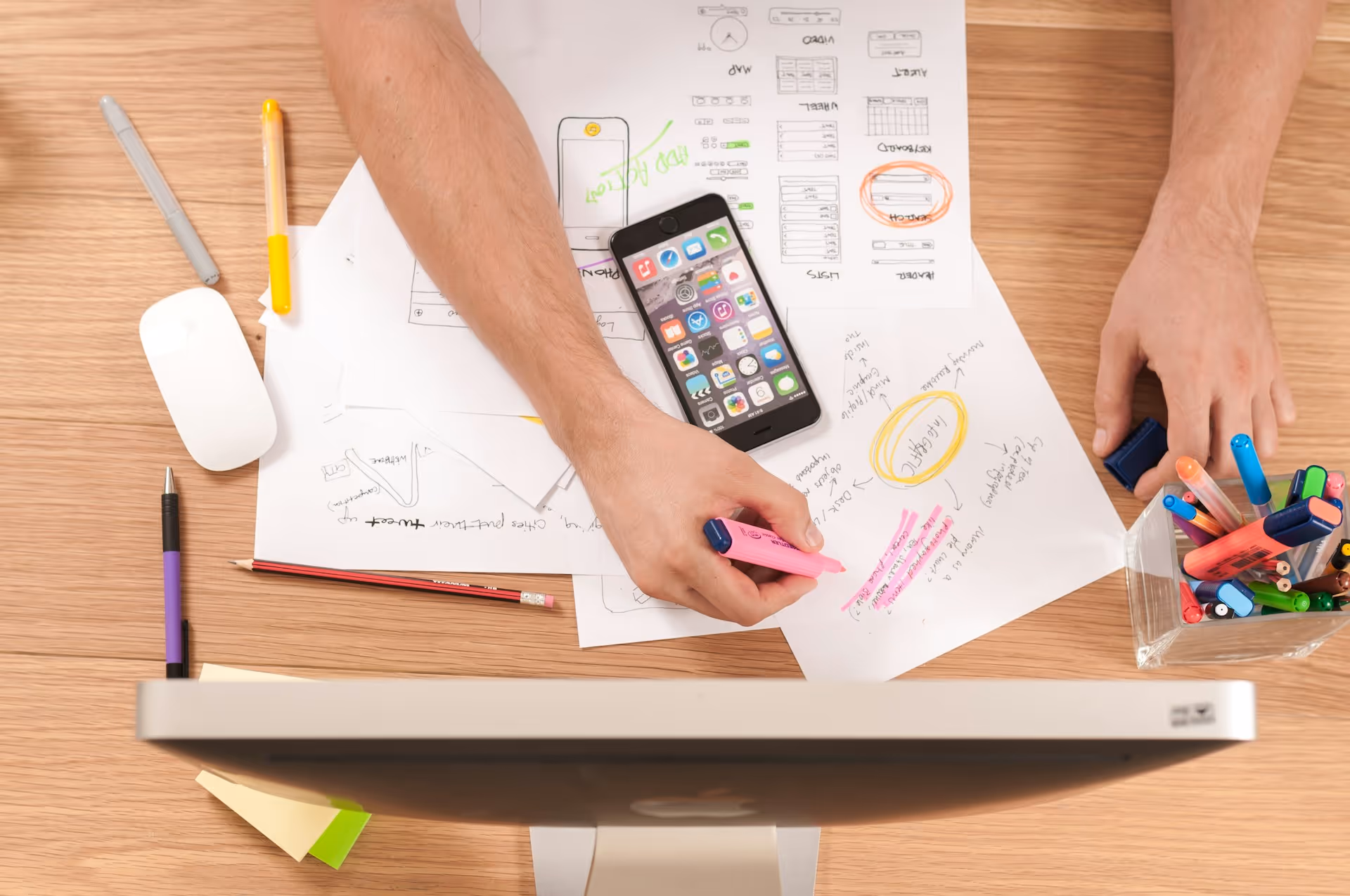 Person highlighting notes with a pink marker on papers next to a smartphone and a container filled with colorful pens and markers on a wooden desk.